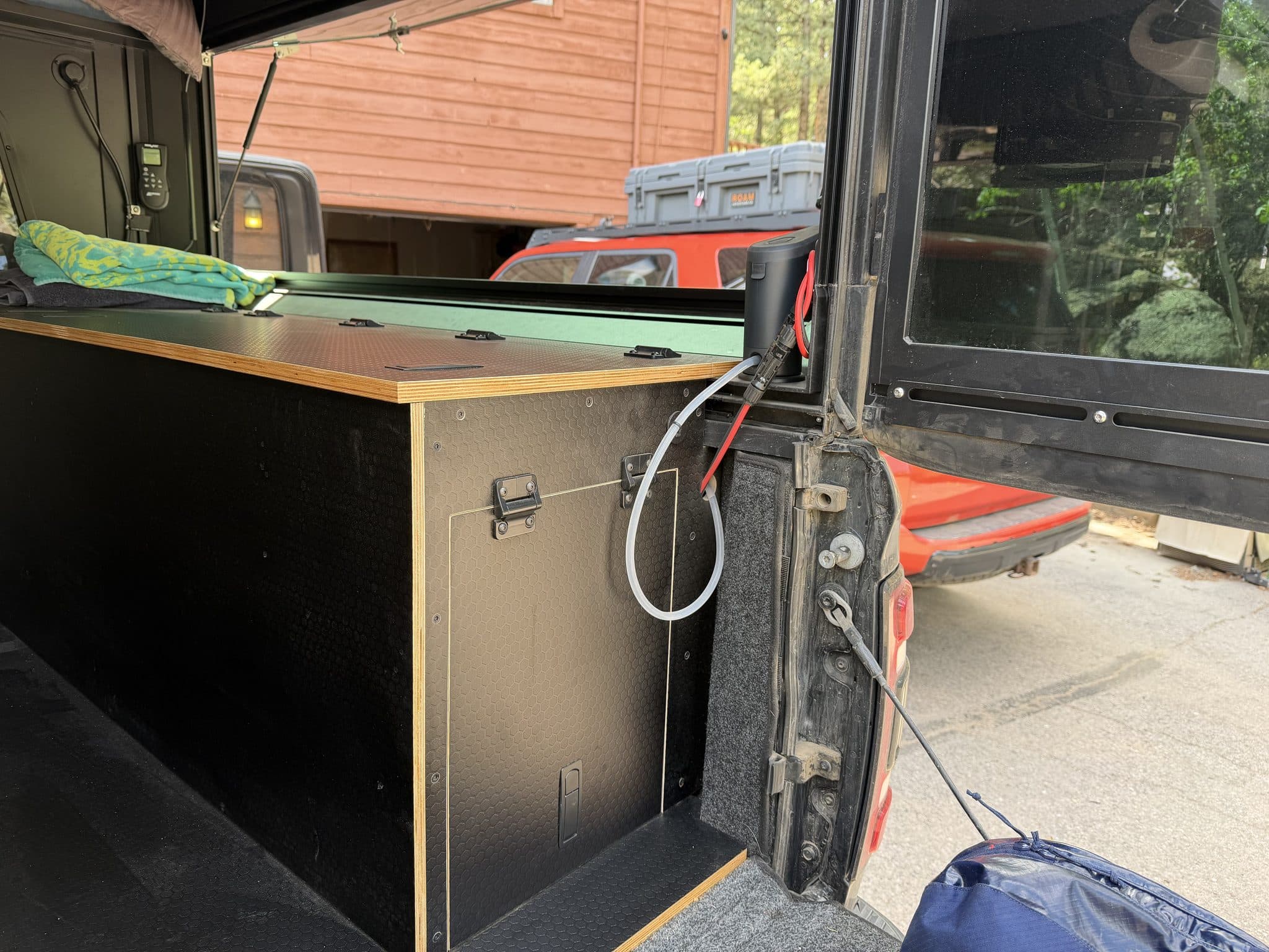 Corner of the plywood cabinet at the open tailgate showing a weatherproof solar input port with an MC4 connector and red wire, next to a latched access panel with stainless hinges.